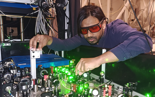 Tahiyat Rahman in a laboratory working with a green laser. Rahman is wearing red safety goggles and a blue shirt.