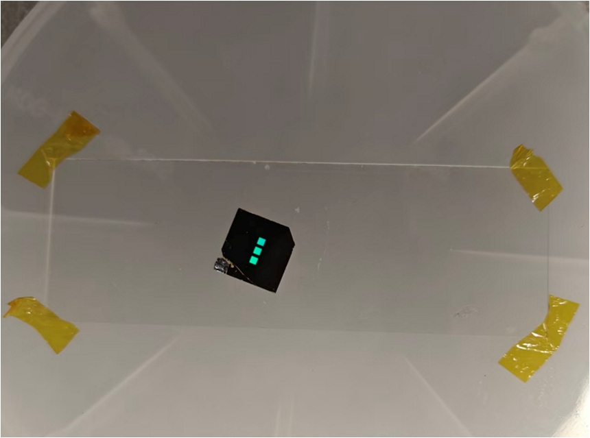 A small, black square metasurface biosensor chip mounted on a clear substrate secured with gold tape, featuring three small vertically aligned squares reflecting a bright turquoise light