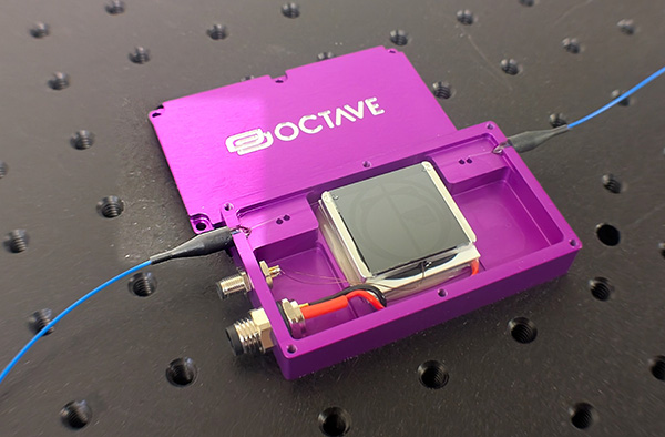 Open purple rectangular casing labeled OCTAVE resting on an optical breadboard, displaying internal photonic components, wiring, power connectors, and connected blue optical fiber cables.