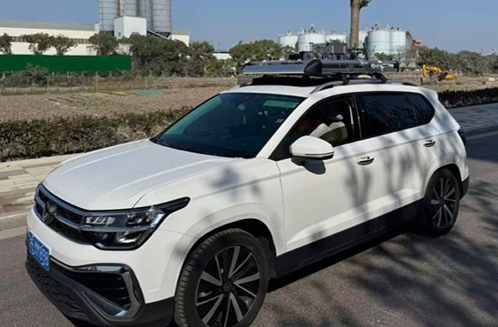 A white SUV equipped with a compact spectroscopy imaging system mounted on its roof rack, driving along an outdoor road near an industrial area.