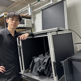 Feng Liu standing in a laboratory, his arm is draped over the QKD setup