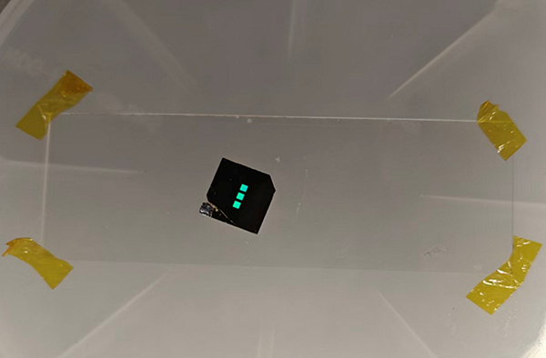 Photo of the chip-based metasurface biosensor, it is a small black chip with three teal-colored lights visible on its surface.