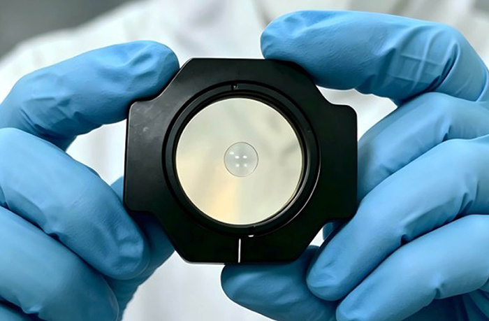 A researcher in blue gloves hold a small, circular, ultrathin diffractive lens housed inside a circular black mounting frame.