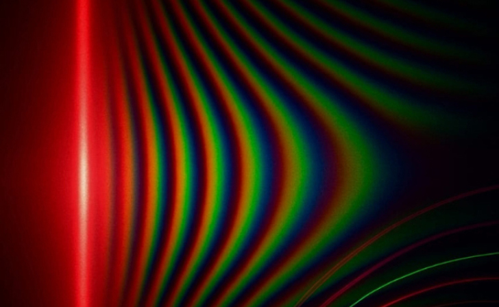 An optical interference pattern showing vibrant, curved spectral bands of red, green, and blue light fanning out across a dark background.