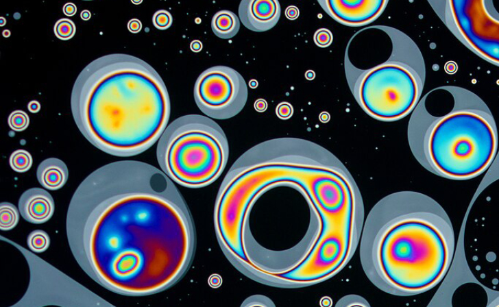 Microscopic view of rainbow-colored, concentric liquid crystal droplets drifting against a black background.