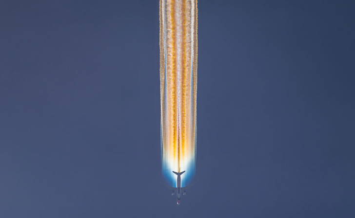 Silhouette of an airplane descending vertically against a dark blue sky, trailing a glowing, iridescent orange and blue contrail.
