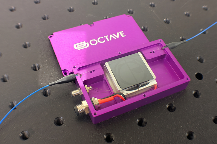 Open purple rectangular casing labeled OCTAVE resting on an optical breadboard, displaying internal photonic components, wiring, power connectors, and connected blue optical fiber cables.