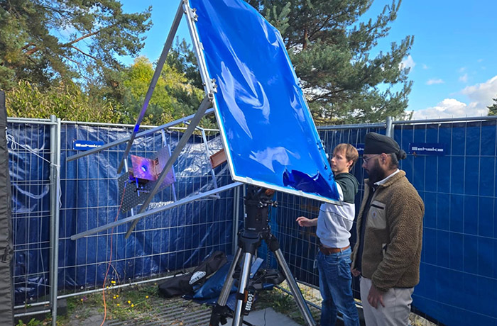 Two researchers standing outdoors examine a large, blue solar concentrator panel mounted on a metal frame and tripod setup.