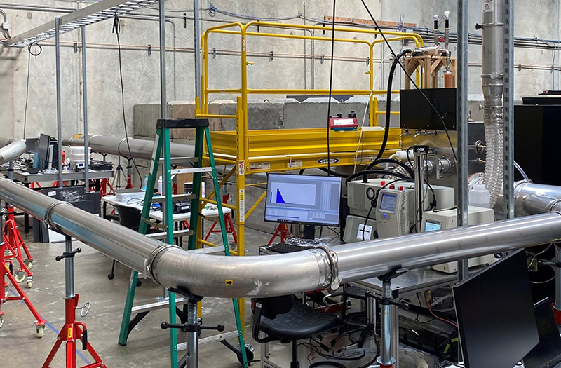 A large laboratory testing facility featuring a long metal aerosol chamber pipe, yellow scaffolding, computer monitoring displaying data graphs, and optical equipment.